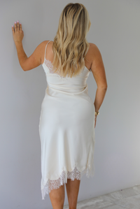 Woman wearing an off white satin midi length dress that has spaghetti straps, lace detailing at the v-neckline, and lace detailing at the hem and split hem detail on the right side. The photo is taken indoors against a white wall and shows the back of the dress.