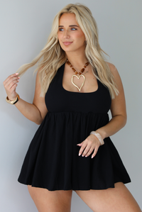 Woman wearing a black romper that looks like a dress, with a halter tie neckline, baby-doll cut, and flowy skirt indoors against a white wall.