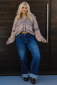 Woman model wearing a mocha brown cardigan that has grey stitching details throughout. The cardigan has a cropped cut, is oversized, and has functional button closures in the front. She's wearing the cardigan with dark denim jeans and brown shoes. The photo is taken against a dark brown wooden door outdoors. 