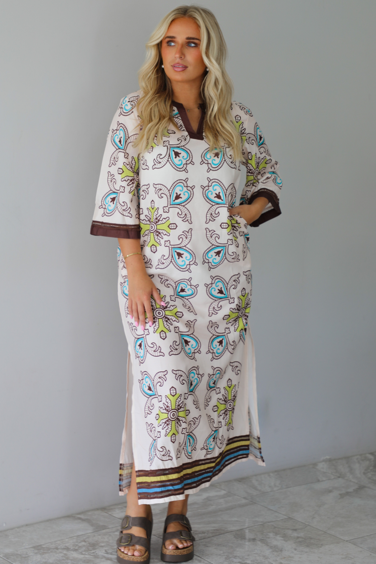 A woman modeling a long kaftan dress with a colorful paisley pattern on a cream background, featuring brown trim and sandals, standing indoors against a white wall with white marble flooring. 