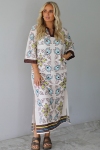 A woman modeling a long kaftan dress with a colorful paisley pattern on a cream background, featuring brown trim and sandals, standing indoors against a white wall with white marble flooring. 