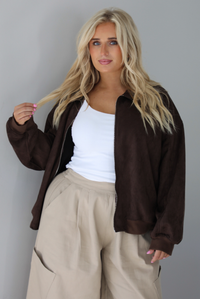 A woman wearing a brown suede jacket with a white tank and sand colored baggy pants, standing indoors against a white wall. The photo shows a closeup of the jacket, which has a zipper closure in the front and cinched details at the wrists. 