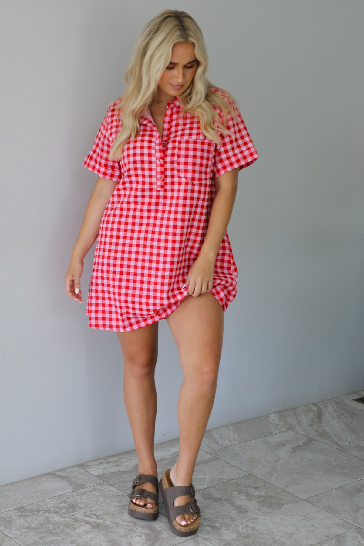 Woman wearing a pink and red gingham collared neckline t-shirt dress indoors against a white wall with white marble flooring. 