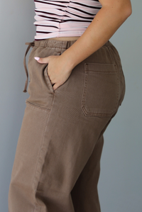 A closeup of a woman wearing mocha barrel pants that have an elastic and drawstring waistline, paired with a pink, burgundy, and white striped design, focused on the pocket on the side. 