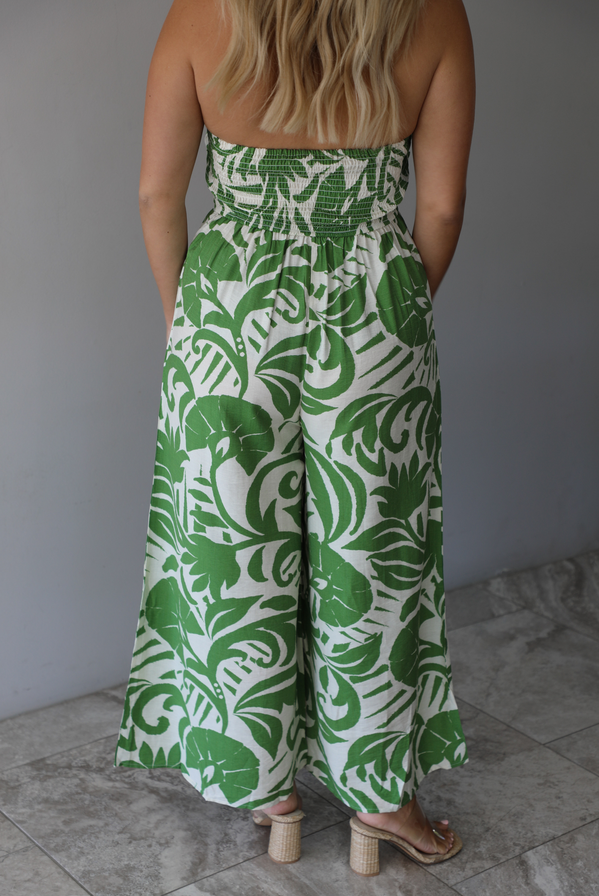 Woman wearing a strapless jumpsuit with green leaf pattern indoors against a white wall with white marble flooring. It shows the back of the jumpsuit, which has a smocked design in the back.