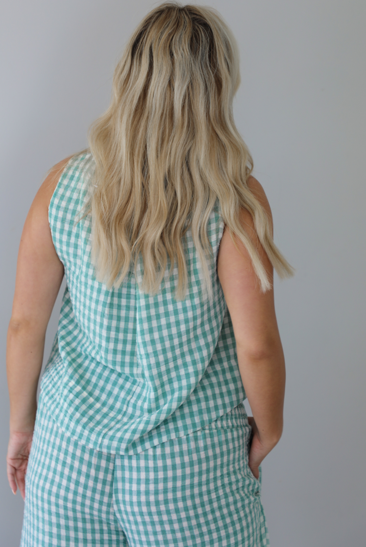 Woman wearing a mint and white gingham set with an oversized top and flared hem pants indoors against a white wall. The photo shows a closeup of the back of the top. 