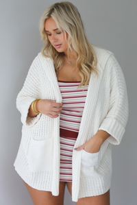 A woman wearing a cream colored oversized cardigan worn over a striped two piece set, standing indoors against a white wall.