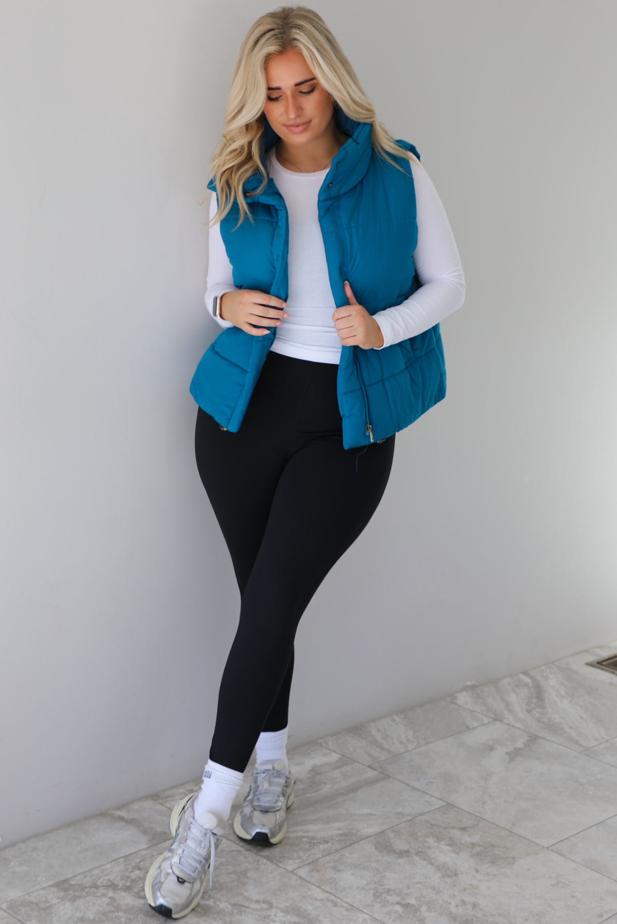 Woman wearing a teal puffer vest, white long-sleeve shirt, black leggings, and gray sneakers with white ankle socks indoors against a white wall with white marble flooring.