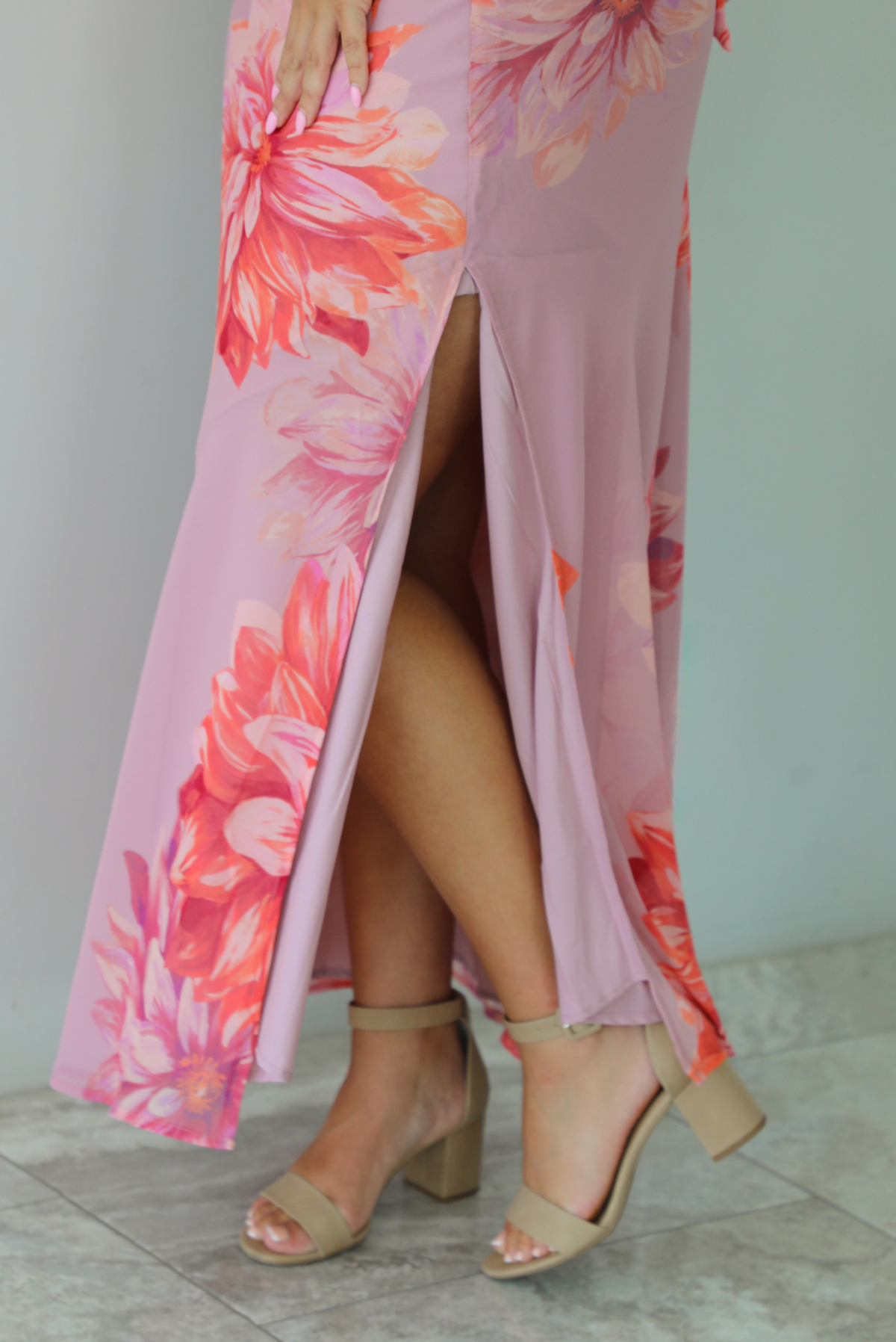 A closeup of a woman modeling a strapless mauve maxi dress with a large red and orange floral print, standing indoors against a white wall with white marble flooring. Th photo shows a closeup of the split hem detail that's on the left side. 