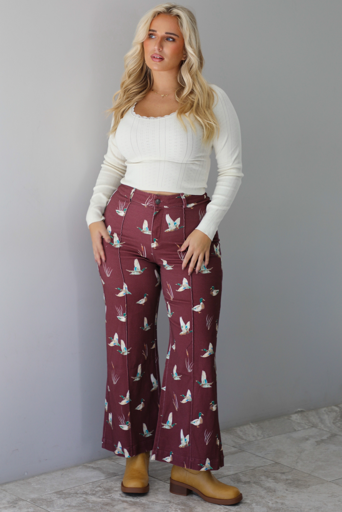 A woman wearing a cream colored knit long sleeved top paired with high waisted maroon jeans with a repeating mallard design, standing indoors against a white wall with white marble flooring. 