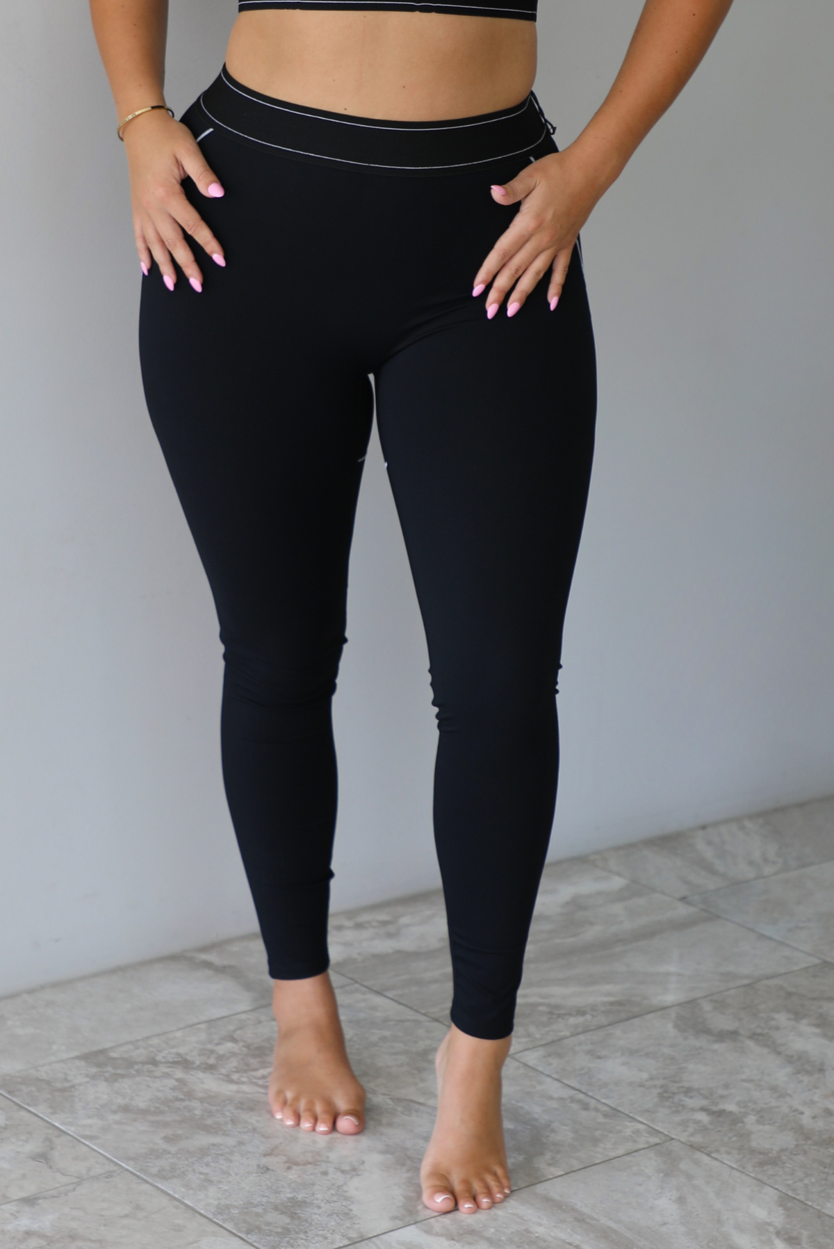 A woman modeling a matching black activewear set consisting of a sports bra and high-waisted leggings with thin white contrast stitching, standing indoors against a white wall with white marble flooring. The photo shows a closeup of the leggings. 