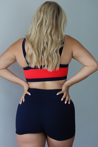 Woman wearing. navy and red sports bra with navy high waisted shorts indoors against a white wall. The photo shows the back of the set. 