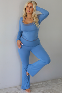 A woman modeling a matching two-piece lounge set in a dusty blue color, featuring a long-sleeve scoop-neck top with a connected darker blue halter neck detail and matching high-waisted flared pants, standing indoors against a white wall with white marble flooring. 