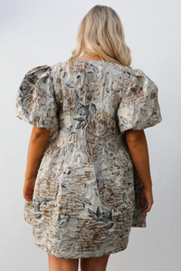 Close up of the back of a woman model wearing a champagne and multi-colored floral pattern throughout. The dress has puffed sleeves, a v-neckline with tie details, and a baby doll cut that stops mid thigh on the 5'7" model. The photo is taken outdoors against a white wall. 