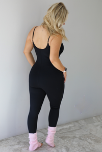 Woman wearing a black fitted jumpsuit with spaghetti straps. The jumpsuit is styled with pink leg warmers. The photo shows the back of the jumpsuit. The photo is taken indoors against a white wall with white marble flooring. 