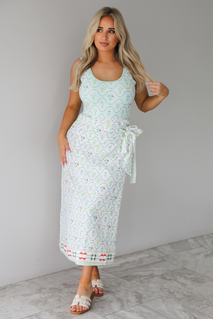 Woman wearing an off white swimsuit & coverup skirt set that has a floral print pattern throughout. The photo is taken indoors against a white wall with white marble flooring. 