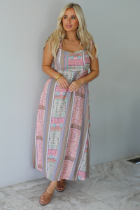A woman wearing a thick strapped maxi dress with a patchwork print of pink gingham, beige paisley, and blue patterns, standing indoors against a white wall with white marble flooring. 