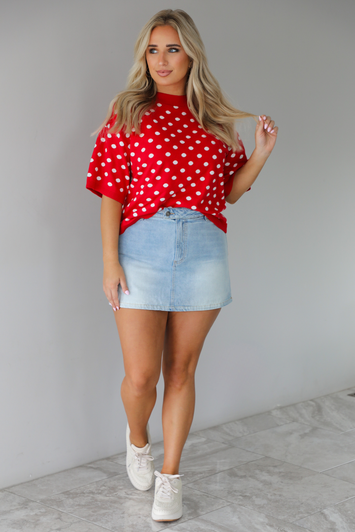 Woman wearing a red knit top with a white polka dot print pattern throughout with a high waisted, light-wash denim skort. The photo is taken indoors against a white wall with white marble flooring. 