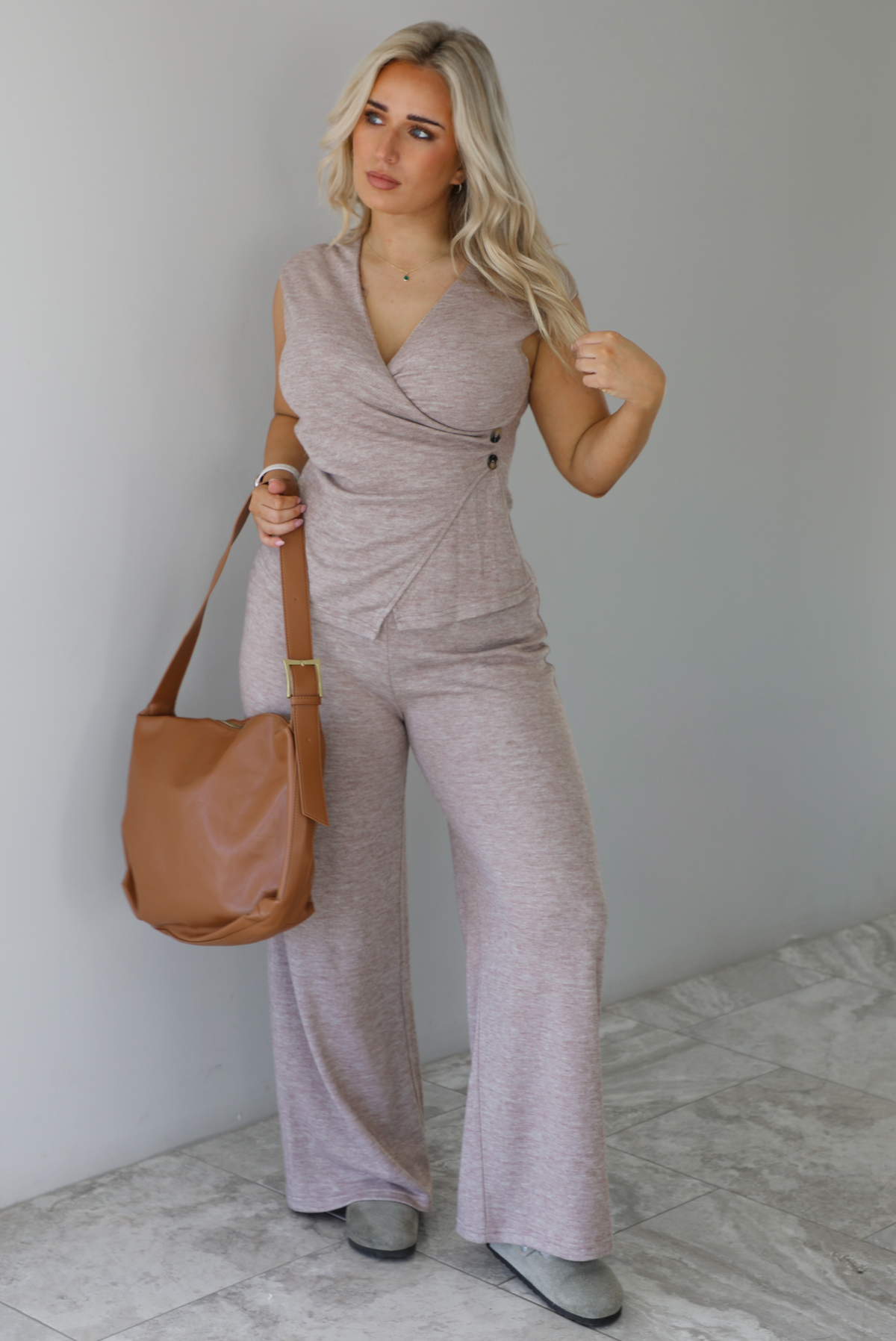 Woman wearing a taupe colored two piece set featuring a vest-appearance top with non-functional button closures on the left side and high waisted pants. It's paired with a tan colored vegan leather shoulder bag. The photo is taken indoors against a white wall with white marble flooring. 