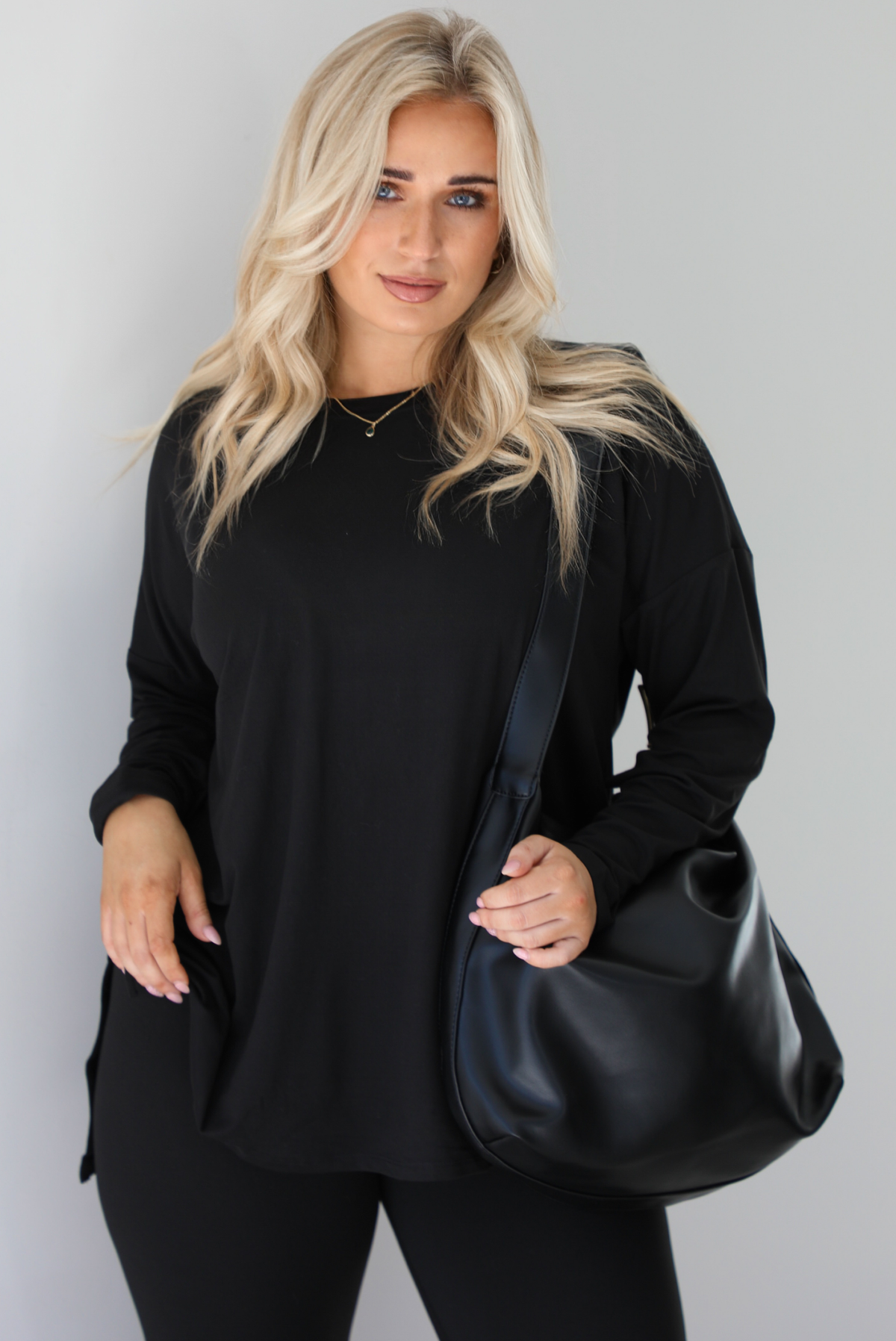 Woman wearing a black two piece set that features an oversized long sleeved top and fitted leggings. The set is paired with a black vegan leather shoulder bag. The photo shows a closeup of the top and bag. The photo is taken indoors against a white wall with white marble flooring.