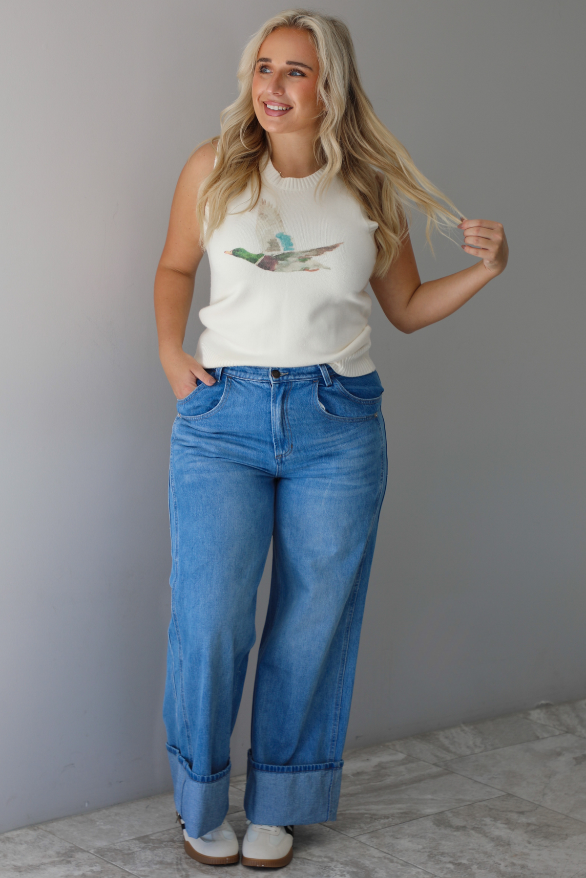 A woman wearing a cream colored sweater vest with a mallard duck on the front, paired with medium wash denim with wide leg hem, standing indoors against a white wall with white marble flooring. 