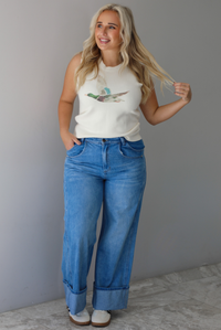 A woman wearing a cream colored sweater vest with a mallard duck on the front, paired with medium wash denim with wide leg hem, standing indoors against a white wall with white marble flooring. 
