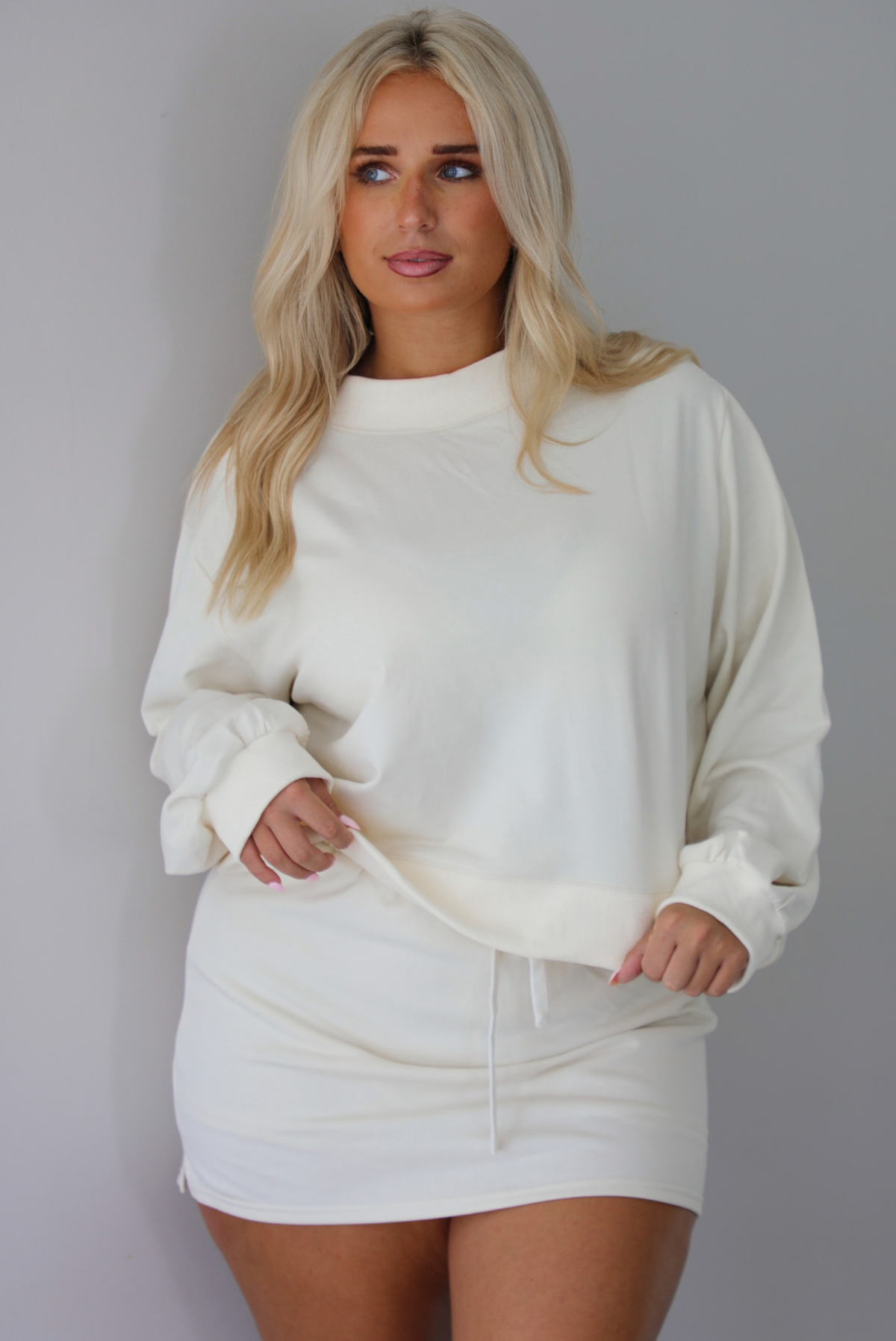 A woman wearing a cream matching two piece set featuring a cropped sweatshirt and mini skirt of the same material, standing indoors against a white wall. The front of the sweater is untucked to show the full length. 