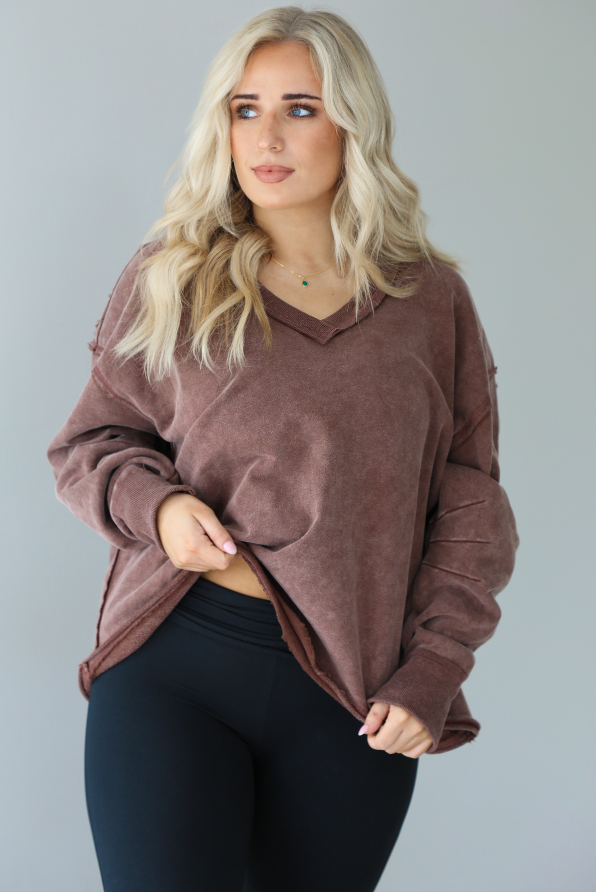 Woman wearing a mauve oversized v-neck sweater with raw hem details with high waisted black flared leggings. The photo shows a closeup of the sweater and the details such as the raw hem and pleated details on the sleeves a the elbow. The photo is taken in front of a white wall.