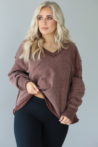 Woman wearing a mauve oversized v-neck sweater with raw hem details with high waisted black flared leggings. The photo shows a closeup of the sweater and the details such as the raw hem and pleated details on the sleeves a the elbow. The photo is taken in front of a white wall.