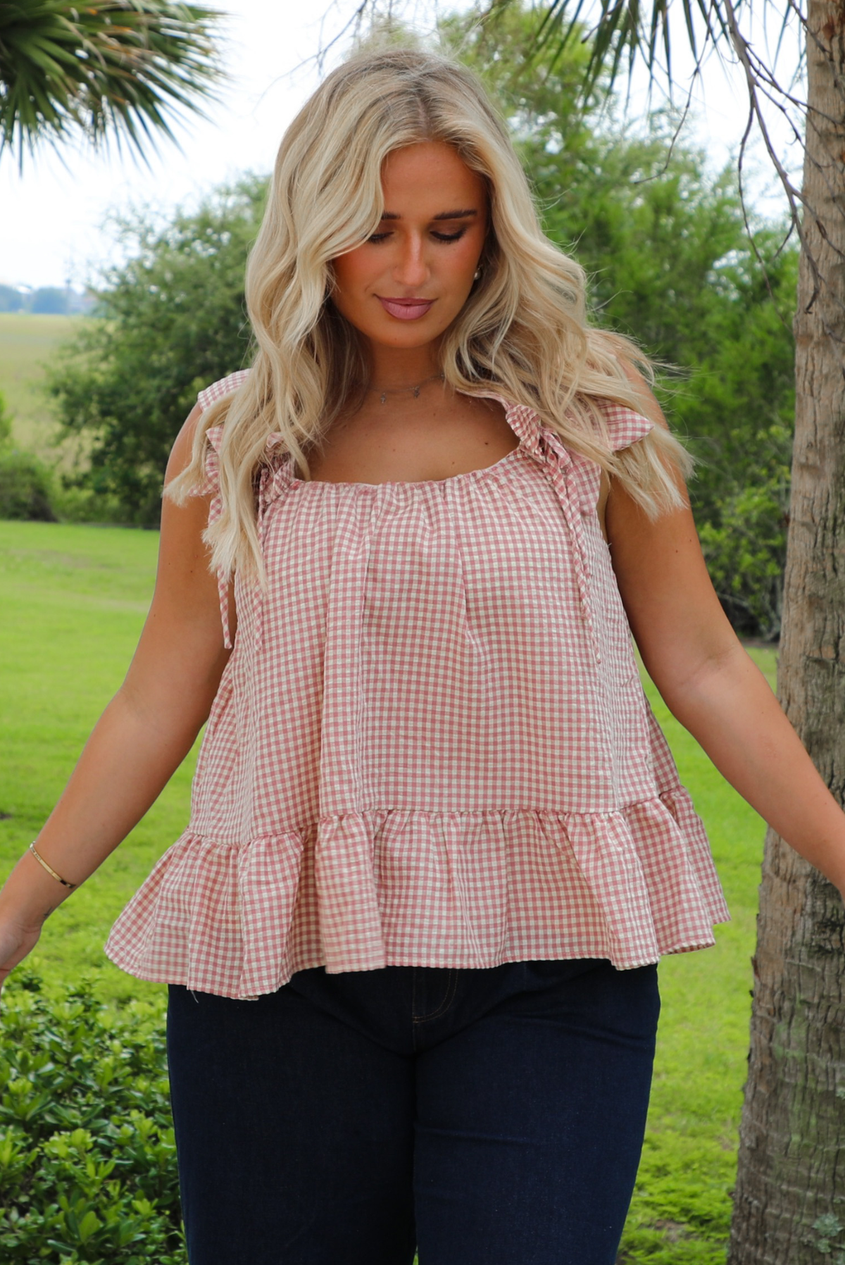 A woman wearing a light maroon and cream gingham flowy top with dark denim jeans, outdoors with greenery in the background.