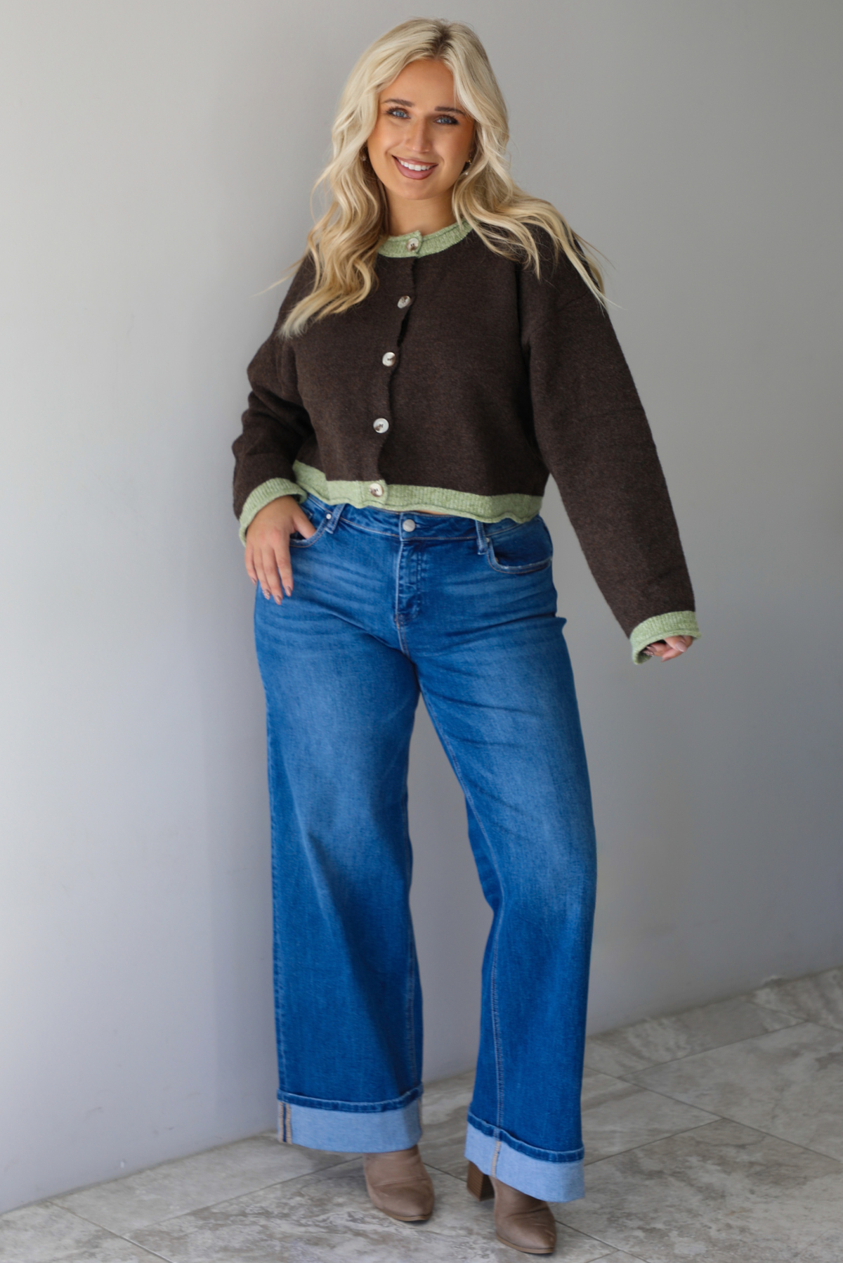A woman wearing a dark brown with green trim cardigan fully buttoned up paired with dark wash, cuffed hem denim jeans, standing indoors against a white wall with white marble flooring. 