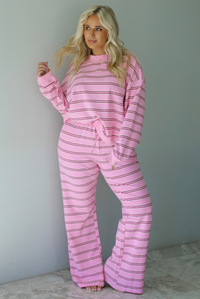 A woman wearing a pink two piece sweat set featuring a maroon and white striped design on a sweatshirt and high waisted, wide leg pants, standing indoors against a white wall with white marble flooring. 