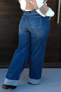 Back of a woman model wearing dark denim blue jeans outdoors. They have a high waistline, functional button and zipper closures in the front, functional front and back pockets, and a slightly flared cut with a cuffed hem. 