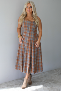 A woman wearing a brown and blue plaid midi dress with thick straps, a square neckline, fitted bodice that starts flaring out at the hips and ends about mid-shin, with a flowy skirt, standing indoors against a white wall with white marble flooring. 