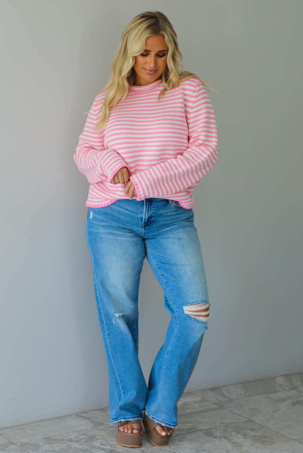 A woman wearing a pink and white striped sweater paired with light wash denim jeans, standing indoors against a white wall with white marble flooring. 
