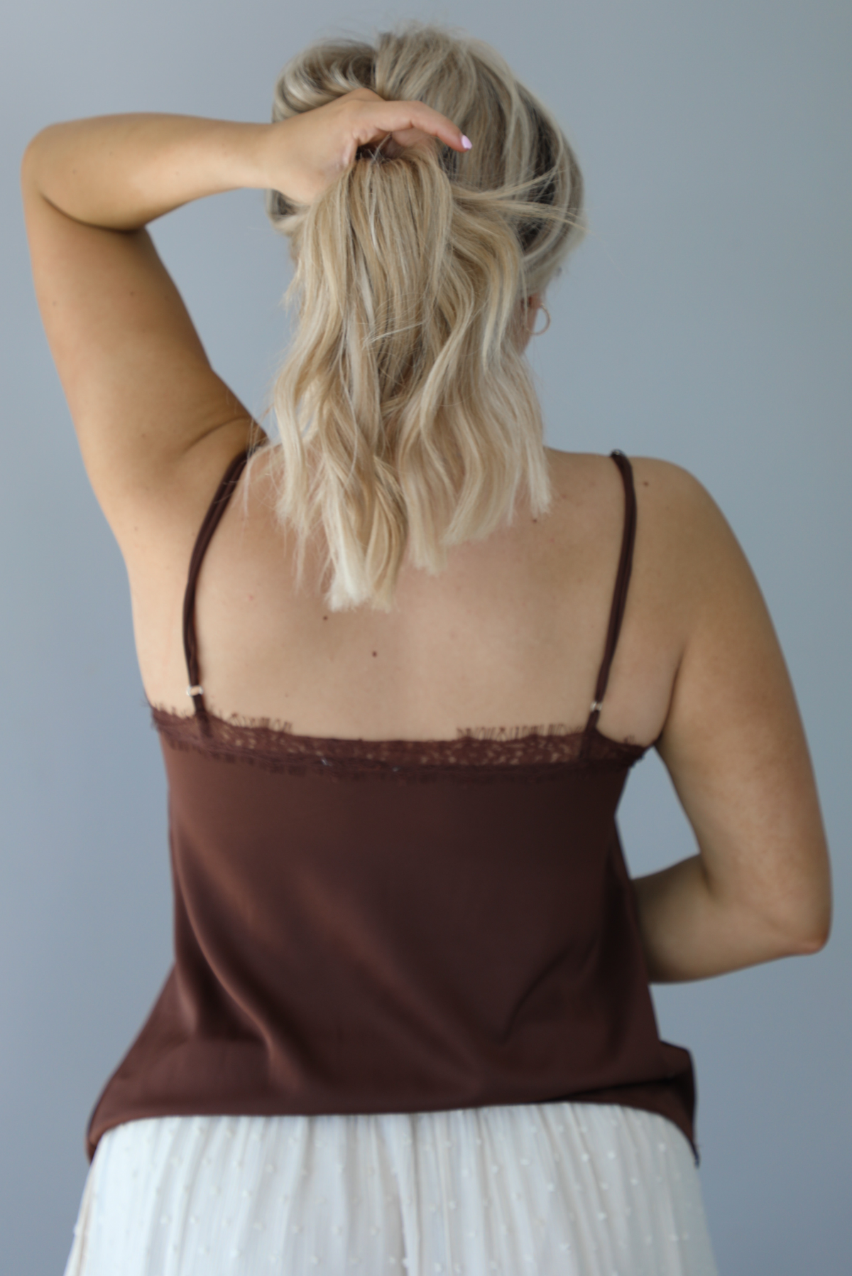 Woman wearing a satin brown tank that is untucked and paired with a high waisted sand colored midi length skirt. The photo is taken in front of a white wall and shows the back of the tank, which has adjustable straps. 