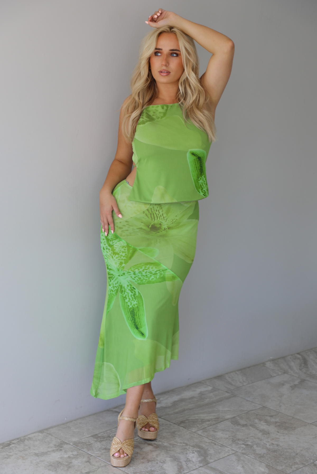 Woman wearing a green tropical print set featuring a backless tank and high waisted, asymmetrical hem midi skirt indoors against a white wall with white marble flooring. 