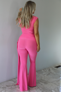 Woman wearing a bright pink jumpsuit that has a high neckline, a fitted bodice, extra fabric detail at the hips with a train on the side, and flared hem pants. The photo is taken indoors against a white wall with white marble flooring and shows the back of the jumpsuit, which has button closures a the very top and a slight cutout underneath.