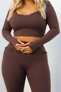 Woman model wearing a brown two-piece athleisure set. The top is cropped and the pants are high waisted. The photo is taken outdoors against a white wall. The photo showcases the top and the thumb hole details on the sleeves, as well as the top half of the pants.