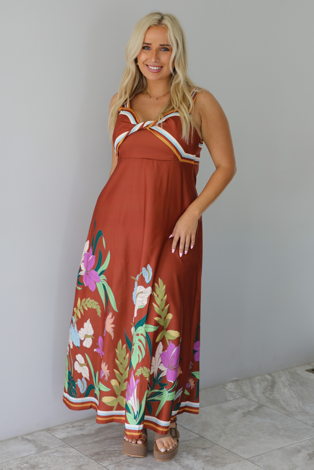 A woman wearing a rust colored maxi dress with other colors at the bust in a striped pattern, with a multi-colored floral pattern at the hem, standing indoors against a white wall with white marble flooring. 