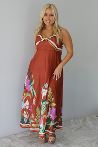 A woman wearing a rust colored maxi dress with other colors at the bust in a striped pattern, with a multi-colored floral pattern at the hem, standing indoors against a white wall with white marble flooring. 