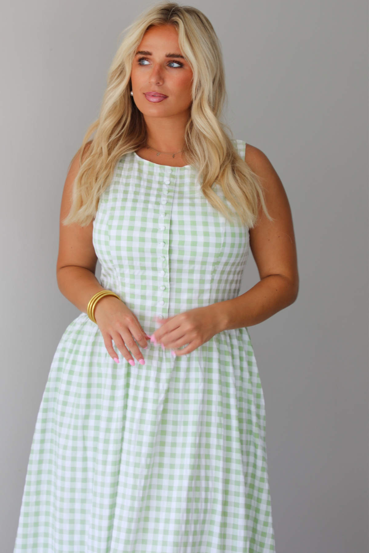 A woman wearing a light green and white gingham maxi dress with a drop waist bodice and side pockets, standing indoors against a white wall. It shows a closeup o the bodice of the dress, which has non-functional button closures in the front. 