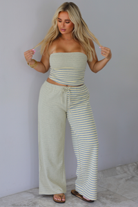 A woman wearing a pale yellow and heather grey striped two piece set with a sleeveless tube top and wide-leg pants, standing indoors against a white wall with white marble flooring. 