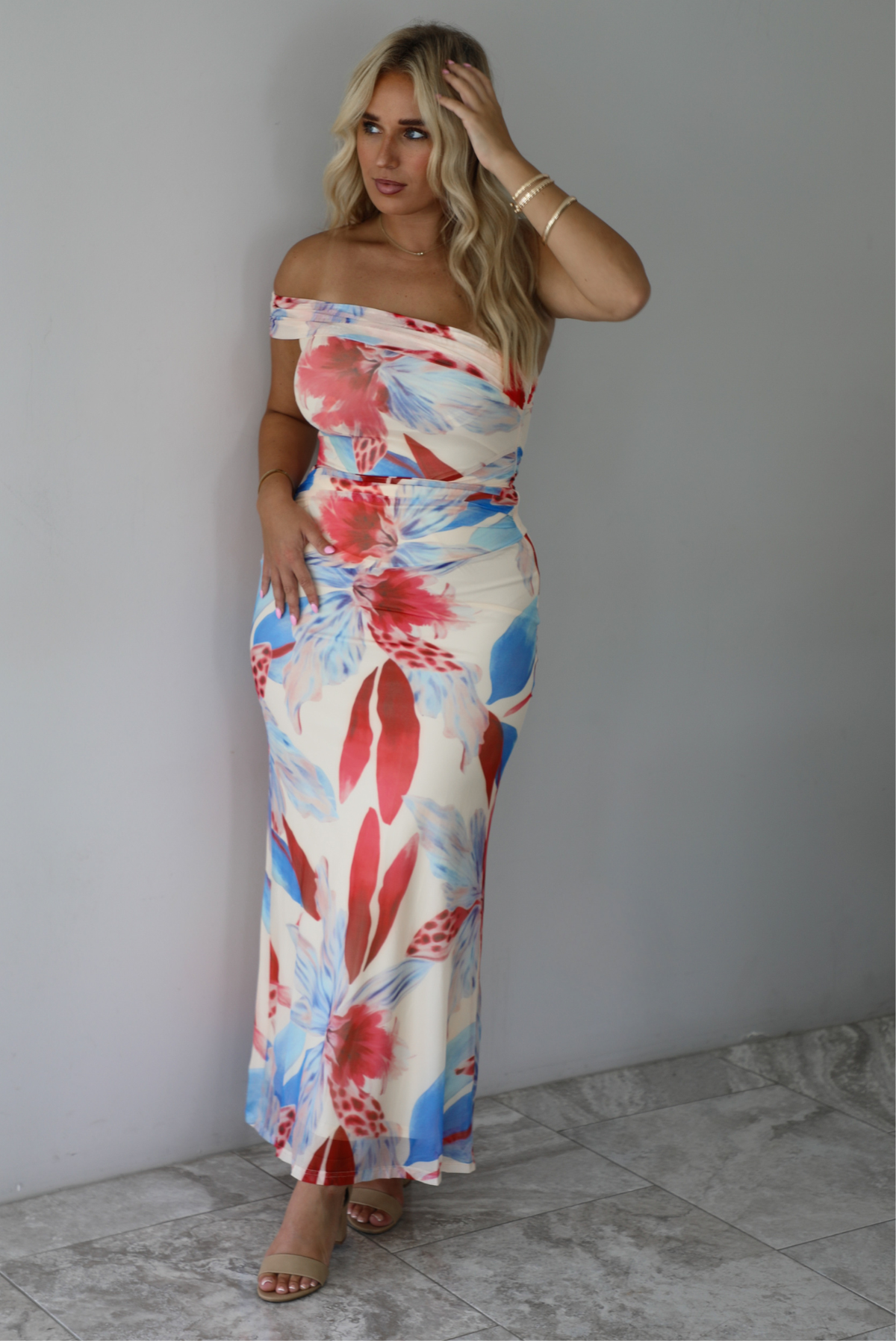 A woman modeling a strapless off-the-shoulder maxi dress with a vibrant red, blue, and cream floral print against a white wall with white marble flooring. 