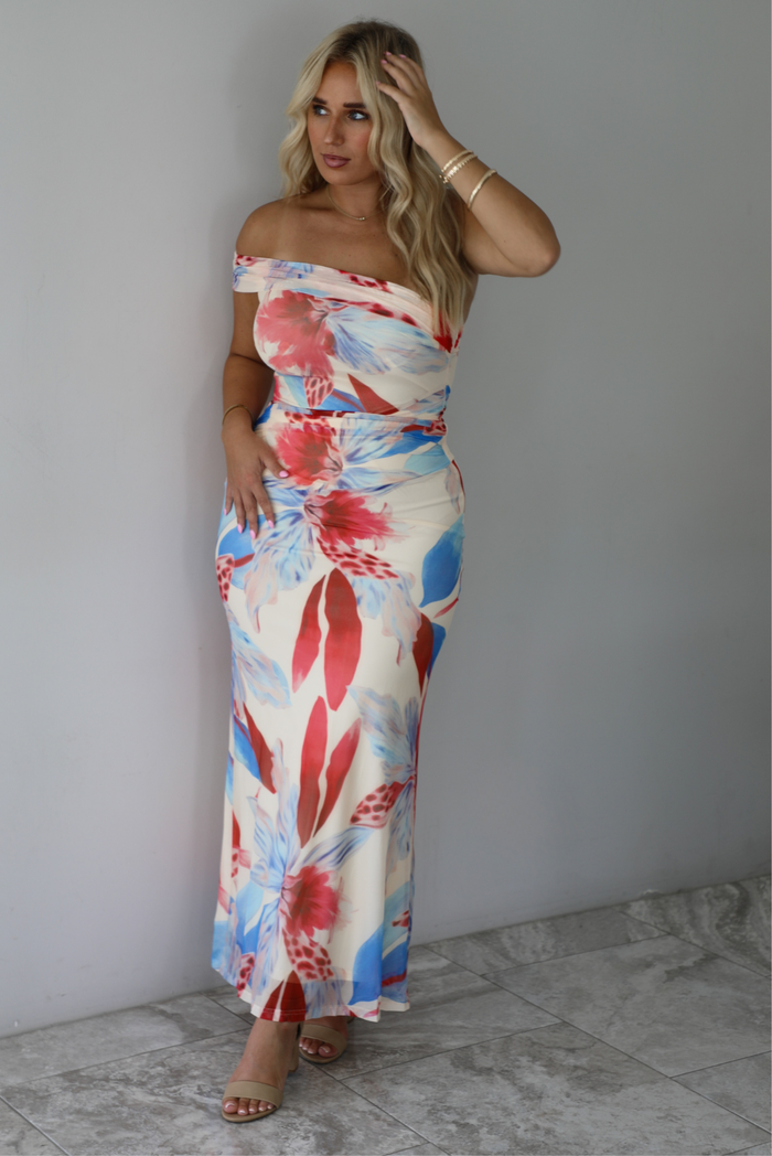 A woman modeling a strapless off-the-shoulder maxi dress with a vibrant red, blue, and cream floral print against a white wall with white marble flooring. 