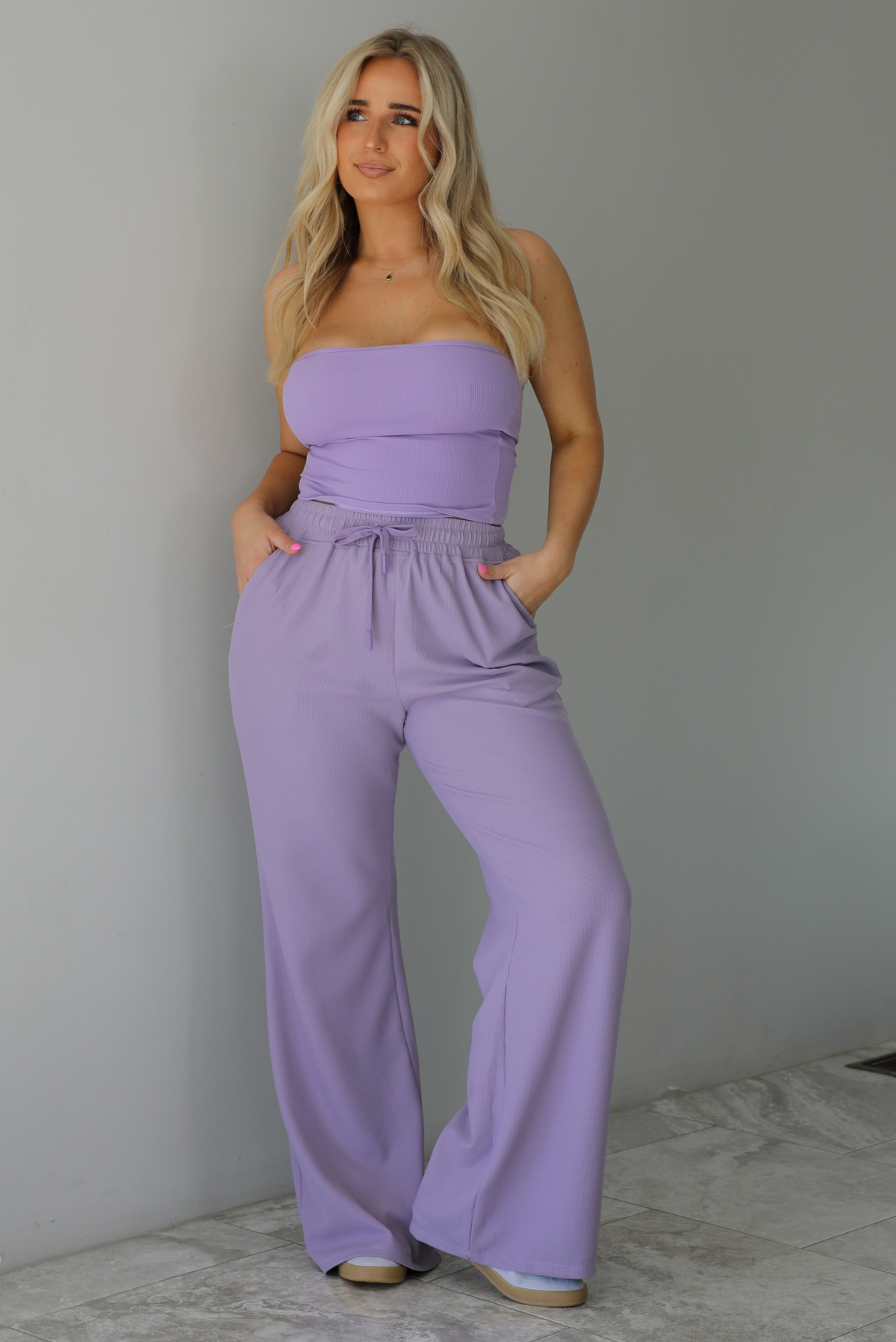 Woman wearing a two piece lavender set featuring a strapless, fitted tube top, and high waisted, flared hem pants. The photo is taken indoors against a white wall with white marble flooring.