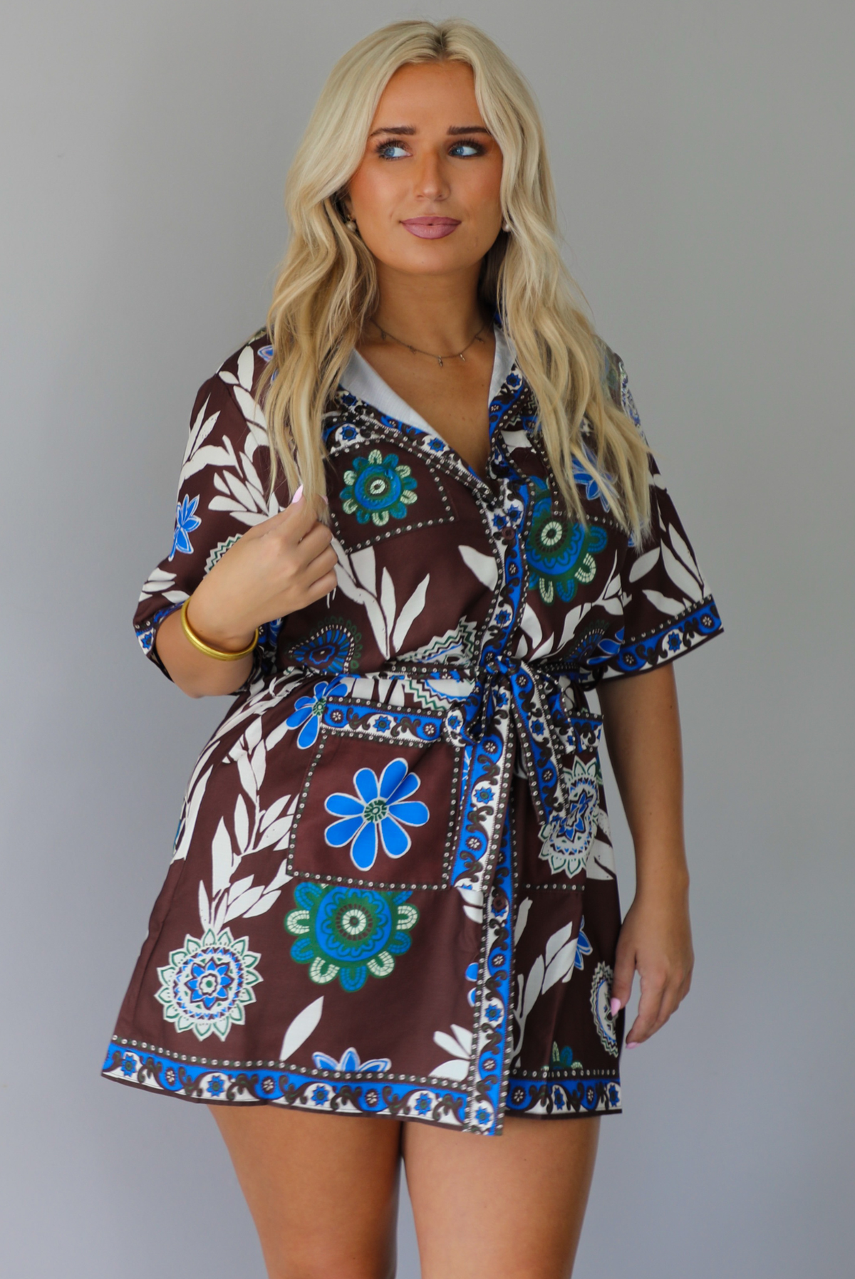 A woman modeling a brown short-sleeve wrap mini dress with a colorful blue, teal, and white floral pattern, paired with beige knee-high boots, standing indoors against a white wall.