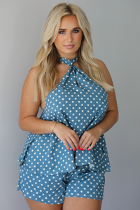 Woman wearing denim blue with white polka dot halter tie top and high waisted shorts indoors against a white wall.