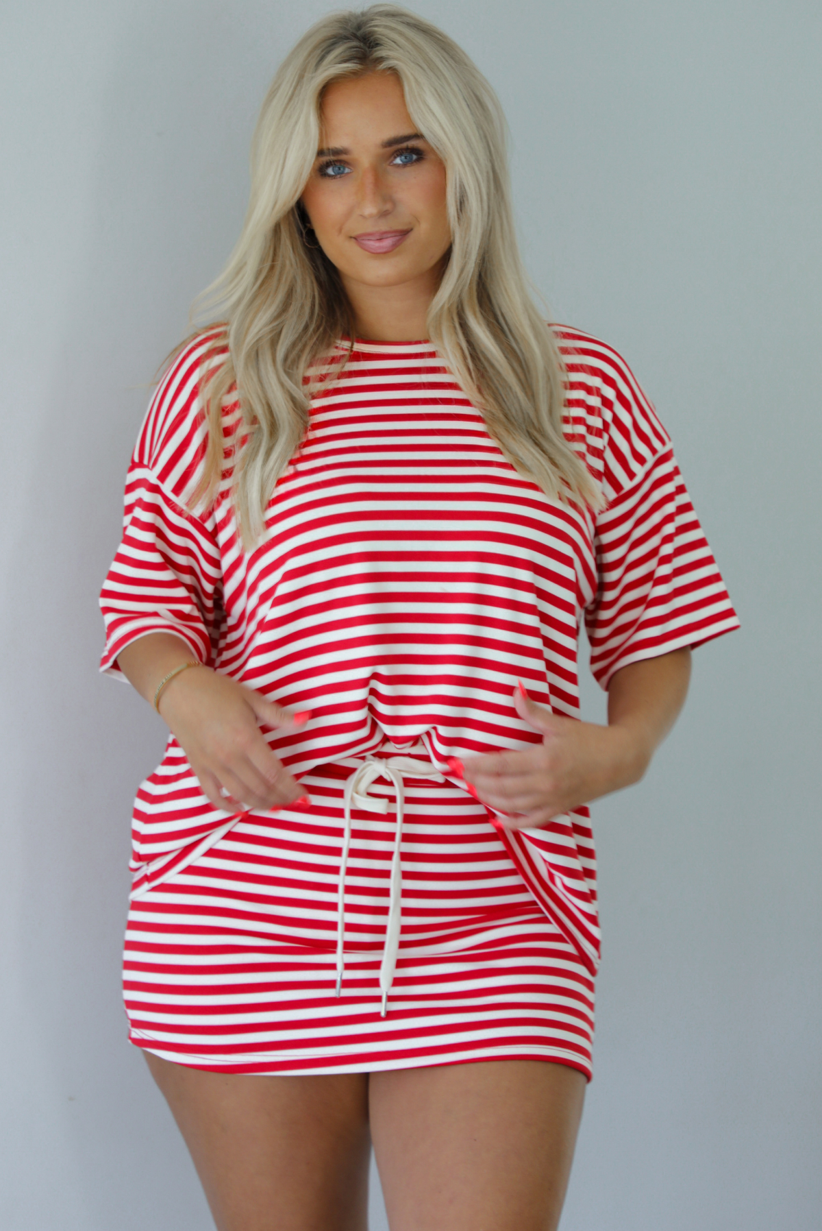 Woman wearing a red and white striped set featuring an oversized top and mini skirt indoors against a white wall. The photo shows a closeup of the set and has the top tucked into the skirt, showing that it has a drawstring waistline. 