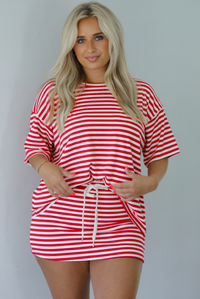 Woman wearing a red and white striped set featuring an oversized top and mini skirt indoors against a white wall. The photo shows a closeup of the set and has the top tucked into the skirt, showing that it has a drawstring waistline. 